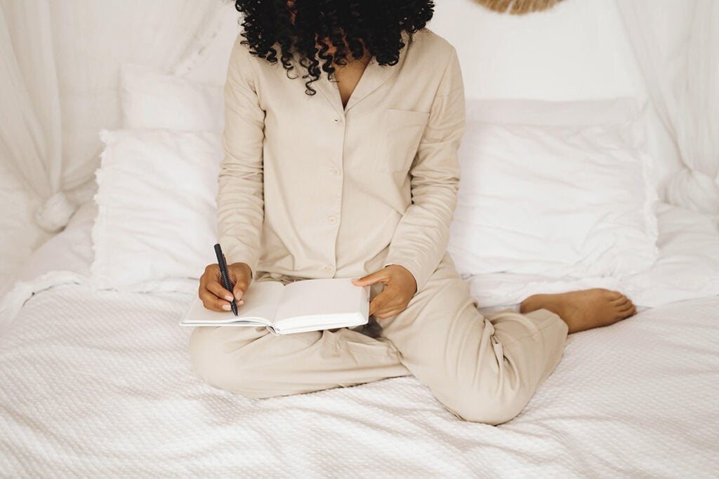 Woman with dark, curly hair writing in a journal on bed